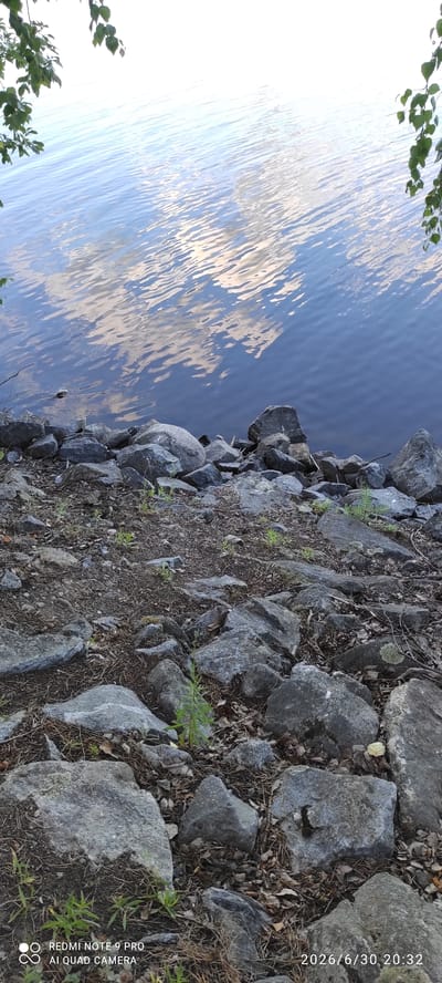 The image shows a rocky shoreline of a body of water, likely a lake, in Hartola, Finland. The water is calm and reflects the sky and clouds, creating shimmering patterns on the surface. There are several large, grey rocks along the shore, interspersed with dry leaves, pine needles, and small green plants. Some of the rocks have lichen growing on them. The scene appears to be outdoors during the daytime, with the lighting suggesting it might be late afternoon or early evening. The overall mood is peaceful and natural.
Visible text on the image indicates "2026/6/30 20:32" and "REDMI NOTE 9 PRO AI QUAD CAMERA", which is a date and time stamp along with camera information. The orientation of the image is rotated 90 degrees clockwise.