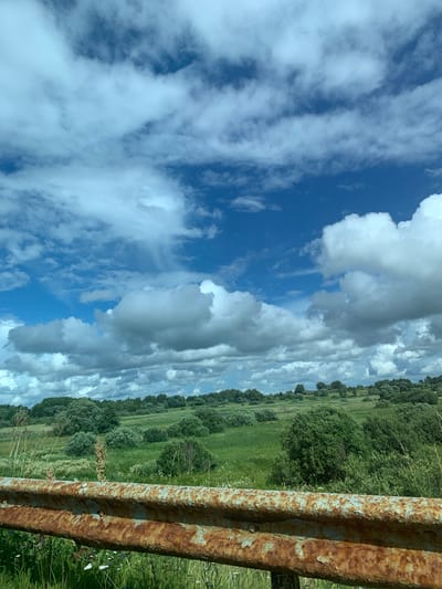 The image depicts a rural landscape under a cloudy sky. In the foreground, a rusty metal guardrail runs horizontally across the frame. Beyond the guardrail, a green field stretches into the distance, dotted with various shrubs and trees. The terrain appears gently rolling. The sky is filled with a mixture of white and gray clouds against a bright blue backdrop. The scene is devoid of people or any discernible activity. Based on the provided information, this image was taken in the Kurapolski Selski Savet, Belarus.