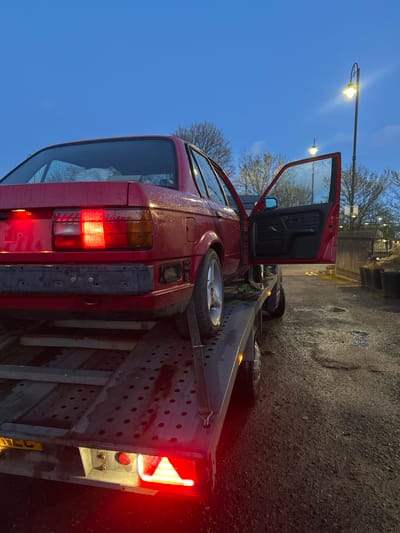 A red car with an open driver's side door is secured to a trailer on a wet asphalt surface. The car is missing its rear bumper. A spare tire is strapped to the trailer near the car's rear. The trailer is parked near a sidewalk adjacent to a fence and some vegetation. A streetlight illuminates the area. The scene appears to be located in an urban setting, potentially in Rutherglen, United Kingdom.