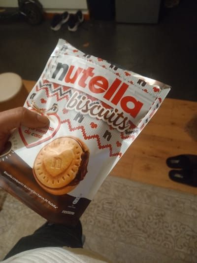 The image shows a package of Nutella biscuits. The package design features the Nutella logo prominently, along with the word "biscuits". A depiction of the biscuit product shows a round cookie with a Nutella filling. Additional text includes "Le vient du coeur" and "Biscuits croquants du coeur onctueux de Nutella." The brand name "FERRERO" is visible in small text on the package. A hand is holding the packet. Background elements include floor, a pair of shoes, and a leg.