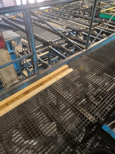 The image shows a close-up, high-angle view of an industrial setting, likely a lumber mill or woodworking facility, in Jēkabpils, Latvia. The primary subjects are various pieces of machinery, including a conveyor belt system with rollers, metal grates, and blue metal railings. Two wooden planks are prominently placed in the foreground, resting on a metal grate. The machinery appears to be in operation or ready for operation, suggested by the organized arrangement of the conveyor belts and rollers.
The scene is brightly lit, possibly by overhead industrial lighting, and there are no indications of weather conditions or time of day beyond the artificial illumination. The location cues are limited to the text "Jēkabpils, Latvia" provided as context, and the overall industrial aesthetic. There are no people visible in the image, nor any discernible emotions conveyed. The focus is on the machinery and the raw materials (wood). Notable details include the dark metal construction of the machinery, the wire mesh of the grates, and the light-colored wood of the planks, some of which appear slightly wet or stained. The image has a utilitarian and functional feel, typical of an industrial environment. There is no visible text within the image itself.