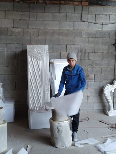The scene depicts an indoor workshop or storage area, located in Andijan, Uzbekistan, characterized by unpainted grey cinder block walls and a concrete floor. A single male individual, appearing young and of Central Asian ethnicity, is positioned center-right. He is dressed in a light grey knit beanie, a blue zippered jacket with white stripes on the sleeves (suggesting an Adidas brand), dark pants, dark gloves, and grey sneakers.
The individual is actively manipulating a large, white sheet of textured foam packaging material, which exhibits motion blur as it is held over a light-colored, two-tiered cylindrical object that serves as a stand or base. His gaze is directed forward, toward the camera.
In the background, to the left, stands a tall, upright, white decorative panel with intricate carved or molded patterns, including scrollwork and a geometric lattice design. Behind the central figure, a larger white object, possibly wrapped, is partially visible. To the far right, a curvilinear, ornate white furniture component is partially visible. The concrete floor surrounding the central figure has scattered pieces of similar white foam packaging material. A thin wooden beam is present along the top edge of the cinder block wall, and some dark electrical wiring runs horizontally across the upper right section of the wall.