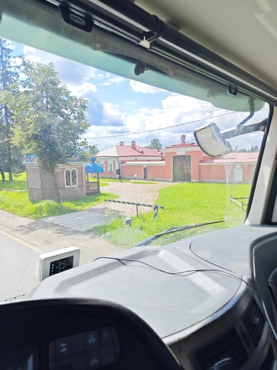 The image is taken from the driver's perspective inside a truck, looking out through the windshield. The scene outside is a sunny day with some clouds. In the foreground, the dashboard and steering wheel of the truck are visible, including a digital display mounted on the dashboard.

The outdoor environment features a road with a yellow line, a grassy area, and a small wooden structure with a blue awning. Beyond that, there's a paved pathway leading to a large building with a red roof and pink walls, featuring a large wooden gate. There's a barrier across the pathway, resembling a railroad crossing barrier. Trees are present on the left side of the image.

The location is identified as Выра (гатчинский), Russia. This suggests the image was captured in or around the village of Выра, within the Gatchinsky District of Russia. The architecture of the buildings and the surrounding landscape are consistent with a rural or historical setting in Russia. There are no people visible in the image.