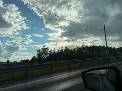 The image captures a view from a vehicle traveling along a road, possibly in or near Lipitsy, Russia. The scene is dominated by a dramatic sky with a mix of bright blue patches and large, gray, cumulus clouds, with sunlight peeking through in the distance. A tall street lamp with two arms and power lines extend across the frame, silhouetted against the sky.
In the middle ground, a row of dense trees forms a dark line along the horizon. Before the trees, a metal guardrail runs parallel to the road, separating it from a grassy embankment. A few houses are visible in the far distance on the left, suggesting a rural or suburban setting.
The foreground shows the wet asphalt of the road, reflecting the light and the sky. In the lower right corner, a car's side mirror is visible, reflecting the road ahead. Inside the mirror, a yellow car is driving in the same direction, further down the road. The overall impression is one of a scenic drive on a cloudy but bright day, possibly during late afternoon or early evening.
