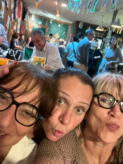 The image displays three adult females in a close-up selfie in the foreground, positioned within an indoor public establishment, likely a bar or pub, in L'Étrat, France.
In the immediate foreground, from left to right:
1. A partially visible adult female with dark hair and black-framed glasses is present on the left. Her right hand, exhibiting red nail polish, is positioned on the head of the central female.
2. A central adult female with dark hair, wide eyes, and puckered lips gazes directly at the camera. She wears a light brown, open-knit garment.
3. A partially visible adult female on the right, with blonde hair and black-framed glasses, also has puckered lips and looks toward the camera. She wears a light brown, open-knit garment and a delicate necklace.
The background shows a populated commercial space characterized by wood-paneled walls, sections painted green, and numerous flags or pennants strung from the ceiling and affixed to walls. A "WC" sign is visible on a green wall, and a dartboard is mounted nearby. Artificial lighting illuminates the area. Multiple adult patrons are present, out of focus. A man with graying hair, wearing a light-colored long-sleeve shirt, is visible in the midground, looking down at a book with a green cover titled "ST PATRICK." Other individuals are standing and conversing throughout the venue, some near tables or a bar counter. A black bag rests on a surface in the left midground, and a yellow bowl is partially visible in the far left foreground. The scene depicts a social gathering inside a public house.