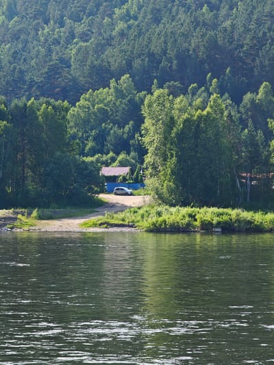 This image depicts a serene waterside scene in Pristan', Russia, with a dense, green forest as the backdrop. In the foreground, the dark, rippling water of a river dominates the lower half of the frame, reflecting the surrounding trees and sky. The riverbank is lined with lush green vegetation and a gravelly shore. Further inland, a dirt road leads to a small clearing where a silver car is parked. Behind the car, a building with a reddish-brown roof is visible, nestled amongst the trees. The forest itself is a thick expanse of various trees, predominantly coniferous, with lighter green deciduous trees interspersed. The overall impression is one of natural beauty and tranquility, with a hint of human presence.