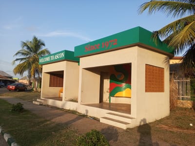 Two concrete bus stops stand side-by-side, each with a flat, green roof. The one on the left reads "WELCOME TO ASCON" in white letters on the green roof. The one on the right reads "Since 1972" in red letters. Both structures are open in the front, with tan-colored walls and steps leading up to the platform. The left bus stop has a single yellow seat. The right bus stop features a mural with green, red, and yellow colors. A decorative, patterned brick window is on the right side of each bus stop. Palm trees frame the scene. In the background on the left, there are parked cars and a building. On the right, there is a white fence and a house partially visible. The foreground consists of grass and a sidewalk. The scene is located in Tawpo, Nigeria.