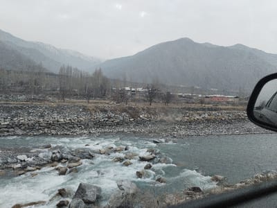 The scene presents a mountainous river landscape under an overcast sky, viewed from the interior of a vehicle. In the foreground, a fast-flowing, greenish-blue river is heavily dotted with large, light grey rocks that create numerous white rapids and turbulence. The river is bordered by a rocky, sparsely vegetated bank. Beyond the bank, a dense cluster of bare deciduous trees stands prominently. Further in the midground, a small settlement is visible, featuring several buildings, including a distinct reddish-pink structure, at the base of towering, grey mountains. The distant mountain peaks are capped with snow. The ambient atmosphere indicates cold, possibly precipitating weather, with fine white specks, consistent with snow or rain, visible across the entire frame. A portion of a car's side mirror, reflecting a light-colored vehicle, is visible on the right side of the image. This scene is located in Qoraqiya, Uzbekistan.