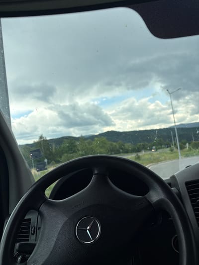 The image depicts a view from the driver's seat of a Mercedes-Benz vehicle, showing the black steering wheel with the Mercedes-Benz emblem and "AIRBAG" text in the foreground. Through the windshield, a landscape under an overcast sky with patches of blue is visible. The scene features green, tree-covered hills or mountains in the distance. A paved road or intersection is discernible, with a tall, light-colored street lamp pole on the right side. Distant, blurry vehicles are present on the road. The windshield shows evidence of minor dirt or water marks. No individuals are visible. The setting is located in the city of Pirdop, Bulgaria.