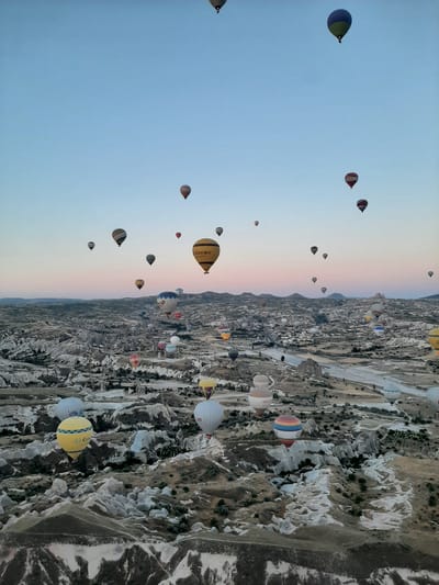 The image depicts a vast, arid landscape in Cappadocia, Turkey, characterized by unique rock formations and valleys. The sky is clear and blue, with a hint of pink on the horizon, suggesting either sunrise or sunset. The main subjects of the image are numerous hot air balloons, which are scattered across the sky at various altitudes. These balloons are of different colors and patterns, with some featuring logos or text, such as "Air Kapadokya" and "UNİVERSAL." The balloons are either ascending or stationary in the air, creating a visually striking scene. The rugged terrain below is dotted with sparse vegetation and some buildings or structures, indicating a sparsely populated area. The overall impression is one of a breathtaking aerial spectacle, capturing the popular tourist activity of hot air ballooning in Cappadocia, a region known for its geological wonders and which includes the city of Avanos.