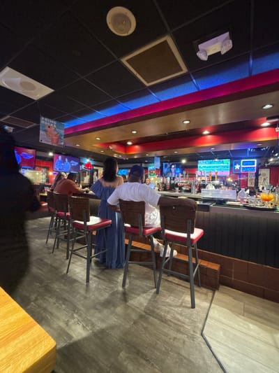 The image shows the interior of a bar or restaurant in Grand Prairie, Texas, USA. The space features a bar counter with patrons seated on red bar stools. Overhead lighting and decorative neon lights create a distinctive ambiance, with multiple television screens displaying content on the back wall behind the bar. Customers are visible at the bar and in the background, interacting with the setting. The floor has a grey finish.
