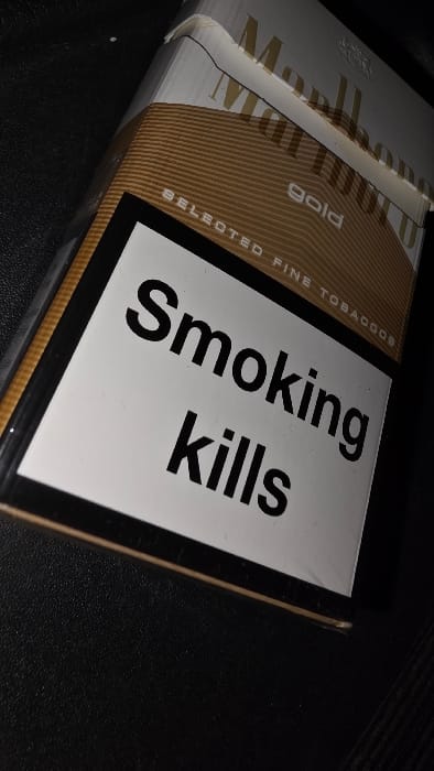 The scene depicts a single, partially opened pack of Marlboro Gold cigarettes positioned diagonally on a dark, textured surface, possibly leather or fabric. The cigarette pack is primarily white and gold/tan, with the brand name "Marlboro Gold" visible. A prominent white warning label with a black border occupies the lower-left portion of the pack, displaying the text "Smoking kills" in bold black sans-serif typeface. The top of the pack shows slight crumpling of the white packaging and the silver-toned foil beneath. The text "SELECTED FINE TOBACCOS" is visible in white letters on the gold section below the Marlboro logo. The environment is dimly lit, with a direct light source highlighting the pack and casting a subtle sheen on the surrounding dark surface. No individuals are present, and no actions or interactions are depicted. This scene is located in the city of Herentals, Belgium.