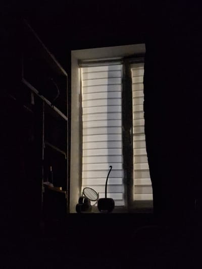 An indoor shot of a dimly lit room shows a window with horizontal blinds, partially illuminated by an unseen light source. On the windowsill, a small, round mirror with a dark frame is propped up, angled slightly towards the viewer. To the left of the mirror are two dark, cylindrical objects. To the right of the mirror, a dark, object with a slender stem curves upwards. To the left of the window, a dark shelf unit with several horizontal bars is visible. The overall scene is dark, with the window area being the brightest part. The city of Apsheronsk, Russia is not visible in this image.