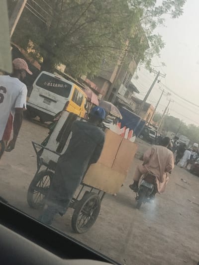The image displays a dusty, unpaved street scene in Gyadi Gyadi Arewa, Nigeria, observed from inside a vehicle. In the left foreground, a person wearing a white jersey with the number "6" and a pink baseball cap walks away from the viewer. Centrally, a person in dark clothing and a blue baseball cap pushes a four-wheeled cart loaded with a large, light-brown cardboard box featuring colorful, pointed decorations on top, and a vertically oriented white object. To the right, an individual in a light-colored traditional garment rides a motorcycle away from the viewer, with a visible puff of white exhaust smoke from the tailpipe. In the mid-ground, a white panel van is visible with "ALLAHU" written in white lettering on its rear window, alongside a yellow vehicle and multiple market stalls topped with colored umbrellas. The background consists of several low-rise buildings, utility poles with numerous overhead wires, trees with light foliage, and additional pedestrians in the distance, all under a bright, hazy sky.