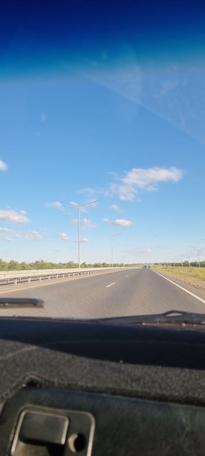 The image captures a view from inside a vehicle traveling on a highway. The road ahead is paved and has lane markings. On the left side of the road, there is a guardrail and a line of trees and vegetation. Several streetlights are visible along the roadside, spaced at regular intervals. The sky is blue with scattered white clouds. In the distance, a few other vehicles can be seen on the highway. The foreground shows the dashboard of the vehicle, including part of the steering wheel and windshield wipers. The location is identified as Bestamak, Kazakhstan. The scene depicts a typical highway environment on a clear day.