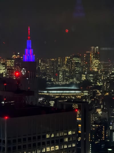 An aerial nighttime view of the Shinjuku district in Tokyo, Japan. In the foreground, the top of a building with large windows and red lights on its edges is visible. Further back, a clock tower is illuminated with a large white clock face. Above the clock tower, a skyscraper is lit with vertical bands of blue and red lights, with a red light at its very top. In the background, a dense cityscape of illuminated buildings stretches across the horizon, with a large, domed stadium or arena structure also visible. The sky is dark and appears to have some faint light reflections.