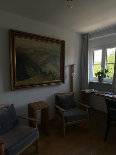 The image depicts an indoor scene, likely a room in a house or hotel, with a prominent landscape painting on the wall. The painting, framed in gold, shows rolling hills and a valley, rendered in muted, natural tones. In the foreground, two upholstered chairs with dark grey cushions and wooden frames are positioned. Between the chairs, a small, slatted wooden side table stands. To the right of the chairs, a tall, minimalist wooden floor lamp with a visible light bulb is placed. Adjacent to the lamp, a white-framed window looks out onto lush green trees. A light-colored curtain is drawn to the left of the window. A wooden desk or shelf runs along the wall beneath the window, with a few items placed on it, including an open book and what appears to be a tablet or notebook. A dark chair is partially visible to the far right of the image, suggesting a desk setup. The overall impression is one of a quiet, slightly dim, and comfortable domestic space, potentially a reading nook or a small study. The provided crops confirm the details of the painting, the furniture, the window view, and the lighting. While the image is set in St. Märgen, Germany, no specific landmarks or features indicative of this location are visible within the frame itself.