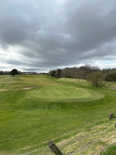 The image shows a view of a golf course on a cloudy day. A yellow flag marks the hole on the green. A sand trap is visible on the fairway. A fence lines the right side of the image, with trees marking the boundary beyond the fence. The scene is likely located in Dunfermline, United Kingdom, based on the general location. Beyond the golf course, the sky is overcast with heavy clouds.