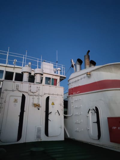 The image displays a detailed view of two white marine vessels or structures, seen from a low angle on a deck against a clear blue sky. The scene is located in the city of Khoroshevo, Russia.

On the left, a white superstructure features multiple windows, likely part of a bridge or upper deck, topped with white railings. Two large, white cylindrical vents with conical caps are prominent. Below the windows, a flat white wall presents two tall, vertically-oriented access panels with dark interiors, a central horizontal grate, and various pipes and conduits. Two yellow electrical junction boxes are present, along with two yellow triangular warning signs: one with a 'T' inside a circle and another with a 'Б' inside a circle. Cyrillic text "АККУМУЛЯТОРНАЯ" (Battery Room) is visible in two locations. A flag with vertical green, white, and red stripes, consistent with the flag of Bulgaria, is partially visible through a window on the upper right side of this structure.

On the right, a separate white vessel or hull section is visible, characterized by a curved form and a bold red horizontal stripe. Above this stripe, three exhaust stacks protrude; two are wide with flared openings, and one is a taller, narrower, rusty pipe. A round, yellow light fixture is mounted on the white surface. Below the red stripe, an elongated black opening with levers is present. A red fire safety box with the Cyrillic inscription "ПР 17" is positioned on the far right.

The foreground shows a dark green deck surface. No people are visible in the scene. The sky is a uniform, deep blue, indicating clear weather.