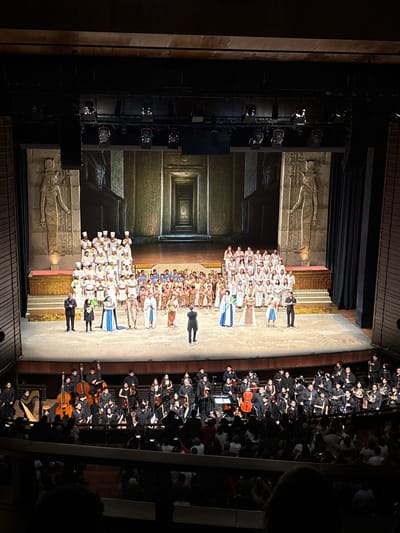 The image depicts a stage performance in what appears to be an auditorium or theater. The backdrop is a digitally projected scene with Egyptian-style imagery, including what appears to be bas-relief carvings and the entrance to a tomb. On the stage are two groups of people, one in the foreground consisting of several rows of people dressed in white hats and what appears to be a white or light-colored gown. In the background, there is an ensemble of musicians in black attire holding instruments. There is a man standing out to the side and is seemingly directing the performance. The lighting is focused on the stage, illuminating the performers and backdrop, while the rest of the auditorium is dimly lit. On the right side of the image, an audience is visible in the seating area. The photograph is likely taken from an elevated position, possibly from a balcony within the auditorium. The location is identified as San Borja, Peru, but the specific venue is not revealed by the photograph.