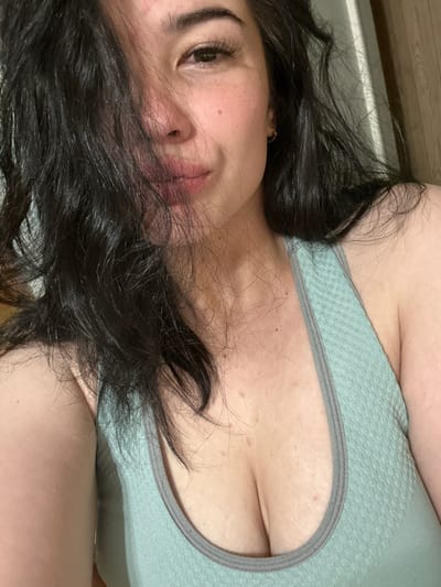A close-up, chest-up, indoor, waist-level shot shows a woman with dark, wavy hair falling across her face. She is wearing a light green sports bra with a gray trim. Her skin appears light-toned with freckles and some redness. The background is blurry but suggests an indoor setting with a wooden surface on the right. No specific location in Gavrilov Posad, Russia is discernible from the image.