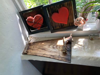 The image displays a windowsill in a room, likely in Negrești, Moldova, illuminated by natural light entering through a window. On the windowsill, two framed red hearts are positioned side-by-side. The left heart is wrapped in wire, and the right heart is plain. Adjacent to the framed hearts is a small white vase containing dried lavender and a crocheted light pink flower. Further to the right, a plant in a white pot is visible, with green leaves reaching towards the window. In the foreground, a wooden serving board with a live edge is resting on a white lace tablecloth. The board is adorned with two deer antlers serving as handles. The overall scene suggests a rustic and decorative arrangement.