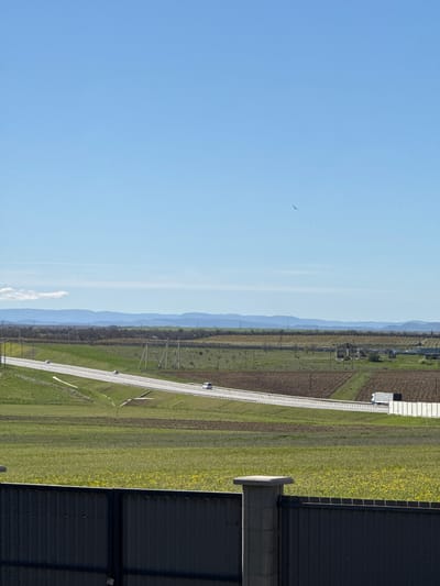 The image depicts a wide, expansive rural landscape under a clear, bright blue sky. The view appears to be taken from an elevated position, possibly overlooking a vast plain.
In the midground, a long, straight road, likely a highway or main rural route, traverses the scene from left to right, receding into the distant horizon. The road features multiple lanes, distinct white markings, and safety barriers or guardrails along its sides. Several vehicles, appearing as small white shapes, are visible driving on the road.
Flanking both sides of the road are extensive agricultural fields. These fields display a mosaic of colors, including patches of tilled brown earth, vibrant green crops, and a prominent section filled with bright yellow flowers, possibly rapeseed or canola.
In the far background, a series of gently rolling hills or mountains extends horizontally, rendered in soft shades of blue and gray due to atmospheric perspective. Along the fields and near the road, several utility poles with power lines are discernible.
On the right side of the foreground, a large building with a dark, corrugated metal roof is partially visible, suggesting an industrial or agricultural structure. In the expansive blue sky, a single bird is seen flying.
The time of day is clearly daytime, with bright sunlight illuminating the scene. The weather is clear and calm, with no significant cloud cover. The overall setting is an open, cultivated countryside, indicative of agricultural activity in the Родниковское сельское поселение region of Ukraine. There is no visible text in the image.