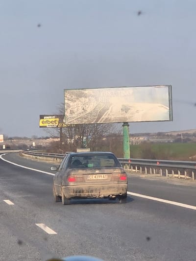 A dark-colored, heavily soiled sedan with Bulgarian license plate CT 4399 AT travels away from the viewer on a multi-lane asphalt highway in Rupkite, Bulgaria. The vehicle's rear bumper and lower body panels are coated in a thick layer of dried mud or dirt. A tow hitch is present below the rear bumper. To the right of the highway, two billboards are visible. The upper, larger billboard displays "ARCUTINO RESIDENCES" and "www.arcutino.com" over an aerial image of a coastal development. Beneath it, a smaller billboard advertises "efbet" with Cyrillic text. A metal guardrail runs along the right side of the highway. The background consists of sparse vegetation, distant hills, and a pale blue sky. The road surface shows white lane markings. No individuals are visible.