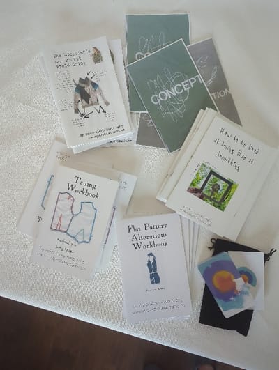 The image displays a collection of instructional booklets and related accessories, consistent with a crafting or sustainable fashion workshop, laid out on a light-colored, textured fabric surface. These materials are associated with Watts Wares, a Philadelphia, United States based initiative.
Specific items include multiple stacks of white-covered booklets. Three identifiable titles, all attributed to "Kimberly Alerts Watts aka Watts Wares" (with website www.wattstwares.com), are "The Upcycler's Pre-Pocket Field Guide," "Sewing Workbook," and "Flat Pattern Alterations Workbook." Another stack of white booklets is titled "How to be Bad at Being Good at Something." Separately, a group of booklets features teal/sage green and grey covers emblazoned with the word "CONCEPT" and abstract line art. In the lower right corner, a small black drawstring pouch holds several small, abstractly designed cards, predominantly featuring circular motifs in orange, blue, and purple. The objects are arranged in distinct groupings across the static indoor scene. No individuals or dynamic actions are depicted.