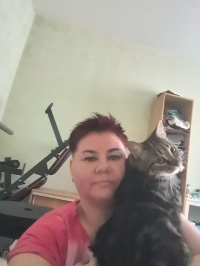A woman with short, reddish-brown hair and an eyebrow piercing is holding a tabby cat in front of her. She is wearing a pink t-shirt. The cat is dark brown and grey with green eyes. Behind them, to the left, is a black exercise machine. To the right is a light brown shelving unit with items on the shelves. The background is a pale green wall. The image appears to be taken indoors, possibly in a home in Verkhnetemernitsky, Russia.