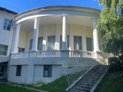 The image shows the exterior of a building with a distinctive curved facade and a portico supported by classical columns. The building is painted in pale yellow and white. There is a white balustrade on the upper level, and a set of concrete stairs with metal railings leads up to the building on the right side. The area around the building is covered in grass, with some trees visible on the right. The sky is clear and blue. The building appears to be in a state of moderate upkeep. This is the building of the "Starorusskaya" sanatorium in Staraya Russa, Russia.