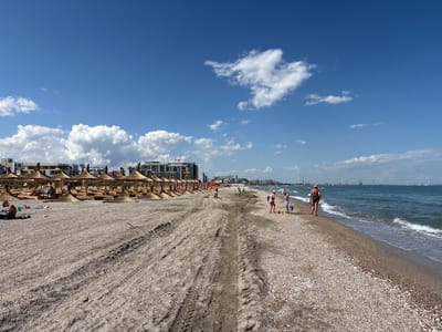 The image depicts a crowded beach in Năvodari, Romania, on a sunny day with a blue sky and scattered white clouds. The beach is covered in a mixture of sand and small shells. Numerous thatched-roof umbrellas and wooden structures are set up in rows on the sand, with some people relaxing under them. In the foreground, tire tracks are visible on the beach, indicating vehicle activity. Several individuals are scattered across the beach. Towards the right, a woman in a green swimsuit stands near the water's edge, facing away from the camera. Further to the left, two young children are playing in the sand with buckets and shovels, accompanied by an adult in a pink hat. In the background, a row of buildings, including apartment complexes and industrial structures, can be seen along the coastline. The sea is a calm blue with gentle waves lapping at the shore.