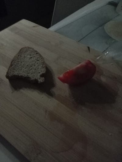 On a wooden cutting board, a slice of dark bread and a wedge of tomato are placed. The background is dark and out of focus, with some bright, indistinct light sources suggesting an indoor environment, possibly a kitchen. The image lacks sufficient detail to determine a specific location within Тулаўскі сельскі Савет, Belarus.