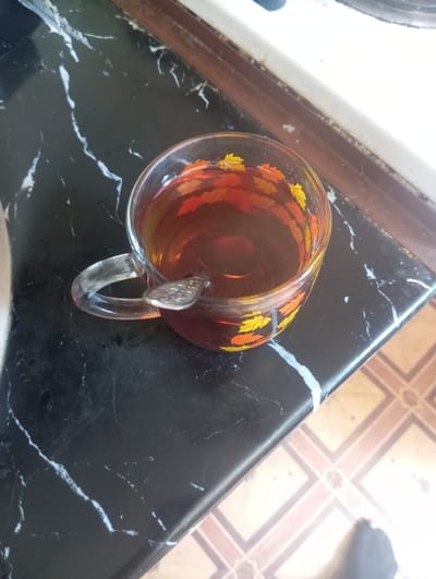 A transparent glass mug filled with dark brown liquid, presumably tea, sits on a dark, marbled countertop. A silver spoon is submerged in the liquid, partially visible. The mug is decorated with a pattern of red and yellow leaves. The background includes a portion of a white appliance, likely a stove, and tiled flooring. The scene is illuminated by natural light. The location is identified as Nurabad, Uzbekistan.