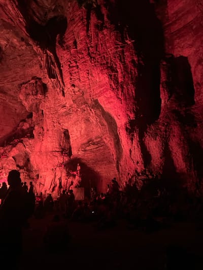 The image depicts the interior of a cave, illuminated by red lighting. Large, rough rock formations dominate the scene, with textured walls and overhangs. In the foreground and mid-ground, a crowd of people is visible, many of whom are seated or kneeling on the cave floor. Some individuals stand in the background, observing the surroundings or interacting with each other. The lighting casts deep shadows, obscuring details of the people and the cave's deeper recesses. The location is identified as a cave in Gollagutta, India, likely a tourist attraction or a place of gathering given the presence of the people.