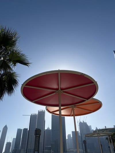 The image shows two large, circular shade structures in Business Bay, United Arab Emirates. The shade on the left is red, and the one on the right is orange. They are supported by metal poles and positioned between a palm tree and a backdrop of numerous high-rise buildings and construction cranes. The sky is a clear, bright blue, with the sun shining through the red shade, causing glare.