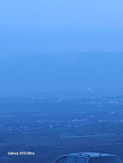 The image displays a panoramic view from an elevated position overlooking a settlement identified as Balsha, Bulgaria, under low-light conditions, likely dusk or dawn, imparting a predominant blue-grey hue to the scene. In the immediate foreground, at the bottom right, the upper portion of a dark-colored vehicle, featuring visible roof rails, is partially present. The text "Galaxy S23 Ultra" is displayed in white font at the bottom left. The midground comprises an expanse of land, likely fields or lower terrain, upon which numerous scattered artificial lights of an urban or semi-urban area are visible. A tall, slender structure, possibly a tower or chimney, is discernible among the illuminated areas in the mid-right section. The background features indistinct, hazy outlines of distant mountains or large hills, softened by atmospheric conditions. The sky occupies the upper portion of the frame, appearing a uniform blue-grey. No individuals, specific actions, or interactions are present within the frame.