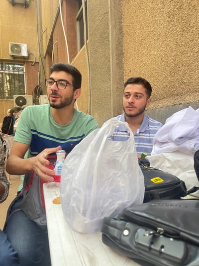 This image depicts two young adult males seated outdoors at a white table in قرية الضبية, Syria.
The male on the left, positioned closer to the camera, has dark hair, a beard, and wears black-framed glasses. He is dressed in a light green t-shirt with a broad dark blue horizontal stripe and dark blue jeans. His right hand is raised and slightly open, positioned near a small white plastic bottle containing a red liquid. A small, light brown, irregularly shaped snack item is visible on the table directly in front of him.
The male on the right has dark hair and a beard, wearing a light blue and white horizontally striped polo shirt. He is looking towards the left side of the frame, away from the camera.
On the white table between the two individuals, a large, clear, crinkled plastic bag stands upright. Further right on the table, two black bags are visible; one features a yellow "CAT" brand logo. A folded white garment is also present on the table, to the right of the plastic bag.
The background consists of a light brown, textured building wall. Two air conditioning units are mounted on this wall, and windows are visible. Distant, blurry figures, including some wearing head coverings, are faintly visible behind the subjects.