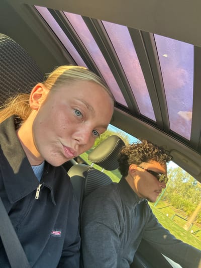 Two people are in a car with a partially covered sunroof. The person in the foreground is a fair-skinned female with blonde hair, and light-colored eyes. She is wearing a dark zip-up sweatshirt. The visible logo on her sweatshirt is a red patch with the word “Dickies” stitched on. She is wearing a seatbelt. Her face is turned towards the camera and she is puckering her lips as if posing for a selfie. The person in the background is a fair-skinned male with curly brown hair. He is wearing sunglasses and a gray zip-up jacket. The male has an earring in his left ear. The scenery visible through the sunroof suggests they are outdoors, likely in a park. Based on the vegetation and sky, it is daytime. The location of the photograph in the city of Serris, France cannot be confirmed based solely on the visual information.