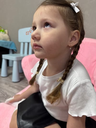 A young girl with light brown hair braided into two pigtails is captured in a close-up shot. She wears a white, short-sleeved ribbed shirt and a black skirt. Her hair is adorned with a small white bow on one side and is tied at the ends of the braids with light-colored bands. The girl has fair skin, light eyes, and a small earring in her left ear. She is seated on a pink cushion. In the background, a light blue child's chair and a plush toy partially covered by a blue cloth are visible. The location is Кипчак, Russia.