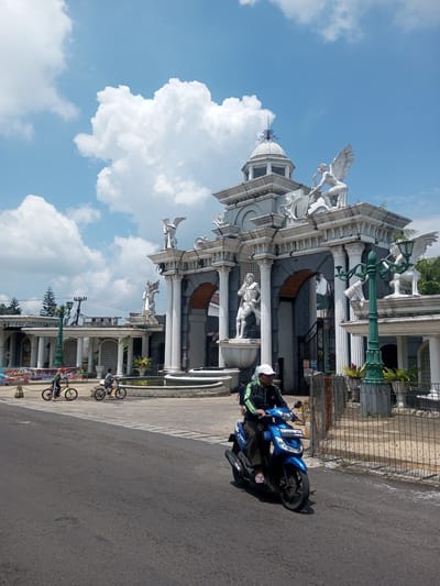 A medium shot depicts a man on a blue scooter in the foreground, with an elaborate architectural structure in the background under a partly cloudy sky. The man is wearing a black jacket, black pants, a white cap, and blue sandals. He is riding on a paved road.
The architectural structure is grey and white, with Roman-style columns, arches, and statues. There are several statues of human figures and winged beings. A decorative dome sits atop the structure. A fountain is in front of the structure. Two people on bicycles are in the distance, near the fountain. A green lamppost is to the right of the frame. A metal fence is on the right side of the road.
The location is an urban area, as indicated by the paved road and surrounding buildings. There is a banner visible in the background. The scene appears to be Xayabury District, Laos.