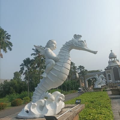 The image displays a large, weathered white statue depicting a winged cherubic figure riding a dynamically posed seahorse, emerging from sculpted waves. The statue is centrally located on a low concrete base in an outdoor, well-maintained landscaped environment under a clear, light blue sky. An active black floodlight is mounted on the statue's base in the immediate foreground. A paved road or pathway curves behind the statue, leading towards a large, ornate archway structure in the mid-ground, partially displaying text that appears to be "SAMUDRA" or "OCEANICA". Another smaller white seahorse statue is visible adjacent to this archway, with a building featuring a domed cupola to its right. The background is populated by numerous tall palm trees and dense green foliage. A bicycle is faintly visible on the road near the archway. This scene is characteristic of Taman Impian Jaya Ancol (Ancol Dreamland), specifically within or near Ocean Dream Samudra or Seaworld Ancol, located in North Jakarta, Special Capital Region of Jakarta, Indonesia.