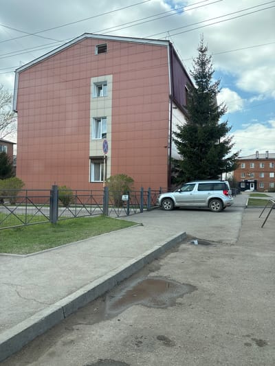 A silver SUV is parked next to a building in Ust-Ordynsky, Russia. The building is multi-story with a tiled facade and multiple windows. There is a tree next to the building and the car. A metal fence separates the building and the sidewalk. The sidewalk is concrete and there is a puddle of water on the pavement. Power lines are visible in the sky.
