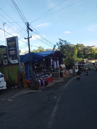 The image depicts a roadside commercial area in the city of Khouathineung, Laos, under a clear blue sky with sparse white clouds. On the far left, a white multi-purpose vehicle is partially parked on a dirt shoulder, adjacent to a low wall painted with green and orange vertical stripes. Above this, a tall black billboard displays text including "SIGNATURE KRETEK," "RP 16.000 BKS," "REVOLUSI KRETEK," and "KOBOY JUNIOR." Central to the left, a dark utility pole heavily laden with numerous overhead electrical and communication wires stands, with several wires extending across the upper frame. Adjacent to this pole is an open-air roadside stall operating under a dark blue tarp roof, supported by metal and possibly wooden poles. The stall visibly displays merchandise, including hanging white and dark-colored garments, multiple stacks of small white packaged goods, and strings of beads or prayer beads, illuminated by several small light bulbs. The asphalt road occupies the mid-ground and right side of the frame, featuring a white dashed line marking the lane. Two individuals, each riding a motorcycle, are observed traveling away from the camera on this road; both riders are wearing dark helmets and clothing. The background consists of a lush, green tree-covered hillside with some scattered distant buildings visible. Further along the road to the right, additional smaller structures or shops are partially visible.