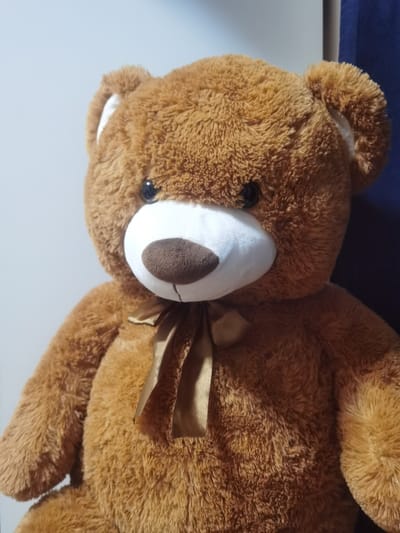The scene presents a large, brown plush teddy bear positioned in an upright, static manner. The bear's fur is a medium brown with a shaggy texture. Its muzzle and the interior of its rounded ears are constructed from white plush fabric. A dark brown, oval-shaped nose is centrally located on the white muzzle, positioned above a subtle stitched mouth line. Two round, black, reflective eyes are set above the muzzle. A gold-colored satin ribbon is tied in a neat bow around the bear's neck. The background consists of a plain, light grey vertical surface occupying the left and central portions of the frame, transitioning to a darker, possibly blue or dark grey, vertical element on the right side. No human subjects, animals, or discernible interactions are present in the image. The visual content does not provide specific identifiers to confirm the requested location in the city of Velichkovo, Bulgaria.