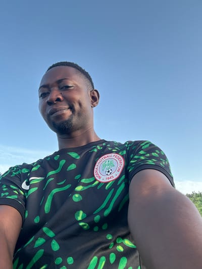 The media file is a selfie of a man wearing a black and green patterned shirt with a circular logo on the chest. The logo has text around it, and a soccer ball is visible inside. The man has dark skin, short hair, and appears to be smiling faintly. He has a small piercing on his nose. The background is a bright blue sky, suggesting it is daytime. The image is oriented sideways. The setting appears to be outdoors, possibly in Gidan Waziri, Nigeria, given the location context. The overall mood seems casual and relaxed.