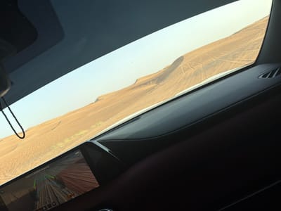 View from inside a car in the desert near Al Madam, United Arab Emirates. Outside the window, there are large sand dunes marked with vehicle tracks. The car's dashboard is visible, along with a rear-view camera display showing the sandy ground. A string of beads hangs from the rear-view mirror.