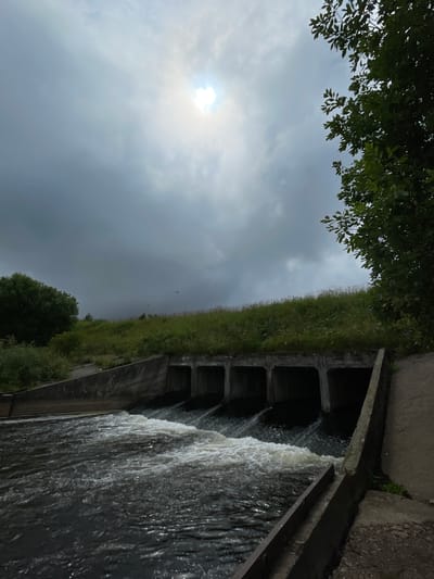 The image shows a spillway or dam structure with multiple rectangular concrete channels where water is flowing through and creating whitewater rapids. The spillway is situated on a grassy embankment with trees visible on the left and right sides of the frame. The sky is overcast with the sun partially visible through the clouds, creating a diffused light. The overall scene appears to be in a rural or semi-natural setting, possibly within the city of Bereznik, Russia, given the context of the query. There are no people visible in the image.