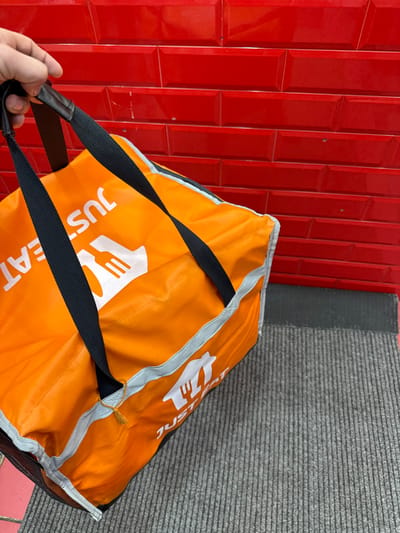 A human right hand, with visible knuckles and short fingernails, grasps the black fabric carrying straps of a bright orange Just Eat insulated food delivery bag. The bag features the white "JUST EAT" logo, including a fork and knife icon, displayed on its visible sides. The background consists of a wall clad in glossy, rectangular red ceramic subway tiles with dark grout lines, showing minor surface scuffs. Below the bag and against the tiled wall, a grey ribbed floor mat is visible, with a narrow dark strip separating it from the wall. A small section of red flooring or tiling is present at the bottom left. The scene is located in Preston, United Kingdom.