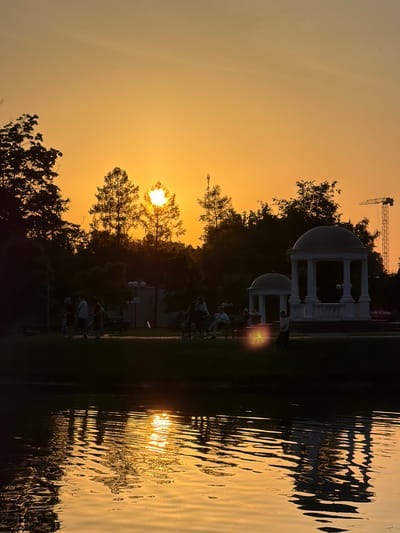 This media file depicts a serene park scene at sunset in Maladzyechna, Belarus. Silhouetted against a warm, orange sky, several people are engaged in recreational activities, suggesting a late afternoon or early evening gathering. The setting includes a body of water reflecting the golden hues of the setting sun, creating a rippling, abstract pattern.
In the background, there are structures that appear to be gazebos or pavilions, adding a touch of architectural interest to the natural landscape. A construction crane is visible in the distance, hinting at ongoing development or infrastructure projects in the area. Trees with lush foliage frame the scene, their branches silhouetted against the bright sky.
The overall mood is peaceful and tranquil, amplified by the soft glow of the sunset. The presence of people suggests a community enjoying the outdoors and the beauty of the evening. The reflection in the water is a prominent detail, showcasing the interplay of light and movement. No visible text is present in the image.
